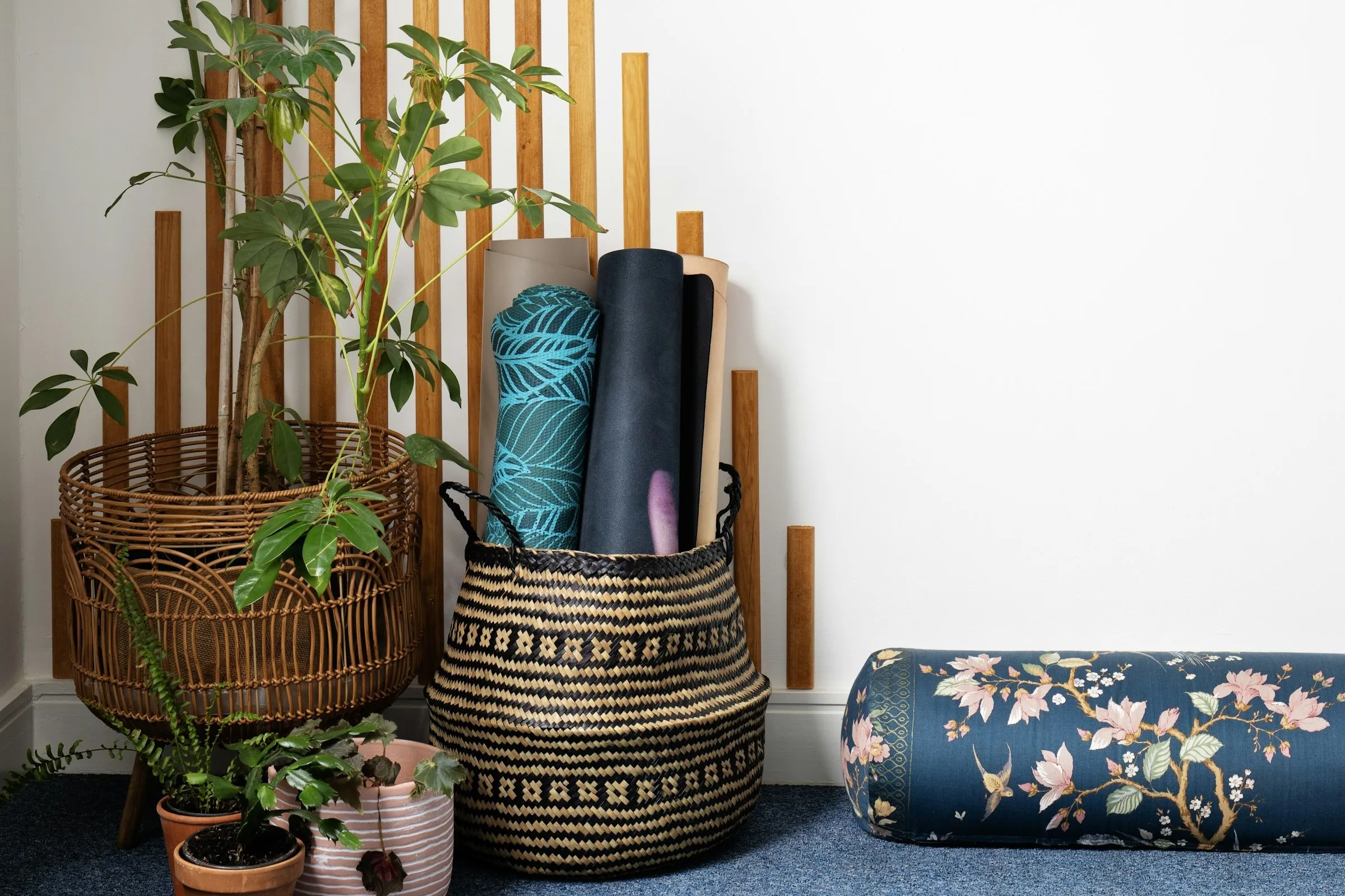 Rolled yoga mats in a woven basket next to a bolster and houseplants in a bright corner
