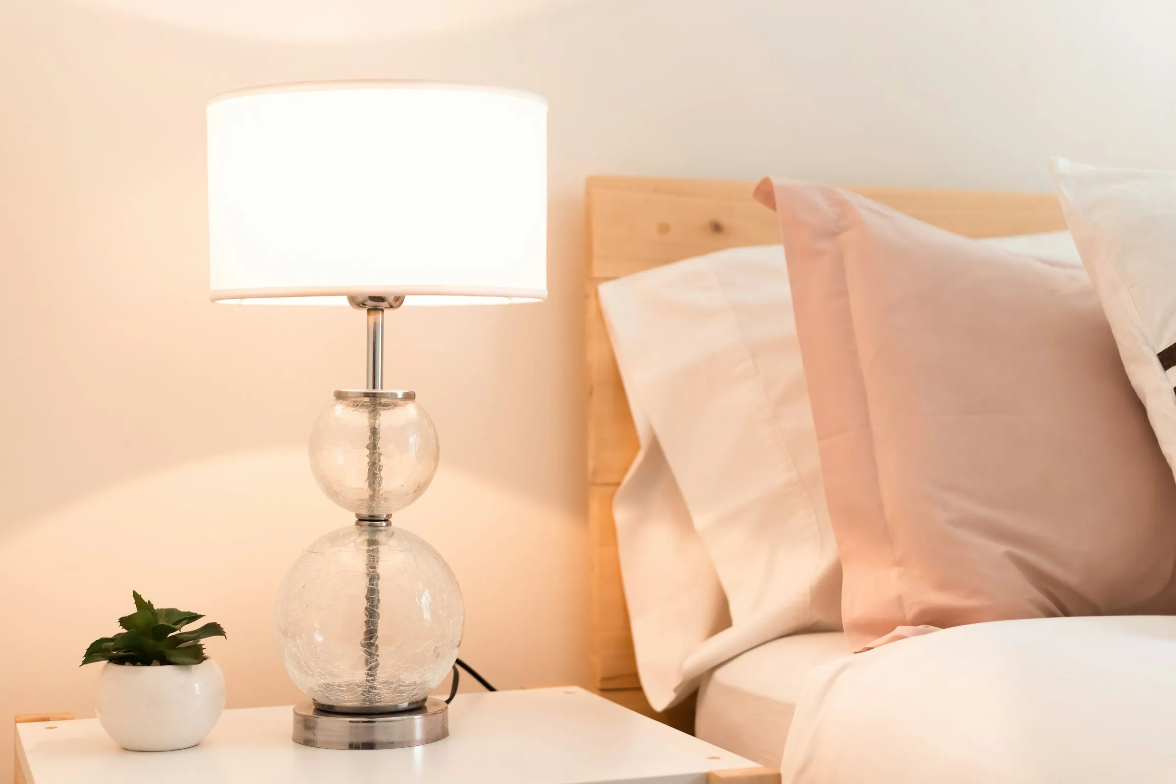 Glass bedside lamp glowing next to pink and white pillows on a bed