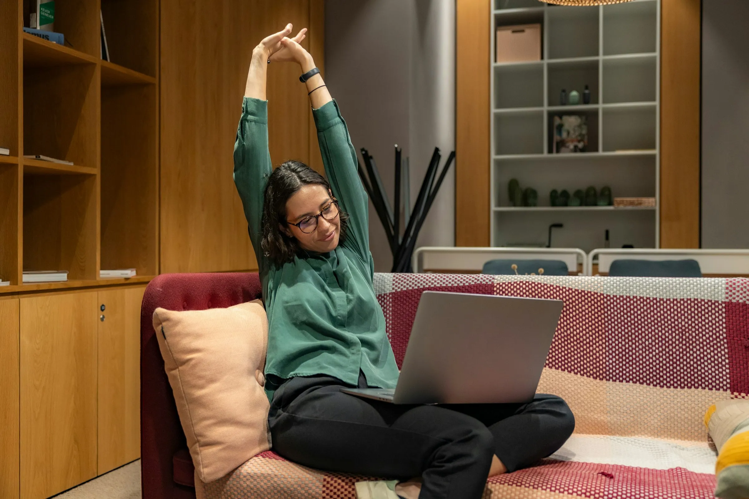 Woman in a green shirt stretching her arms overhead on a sofa with a laptop on her knees