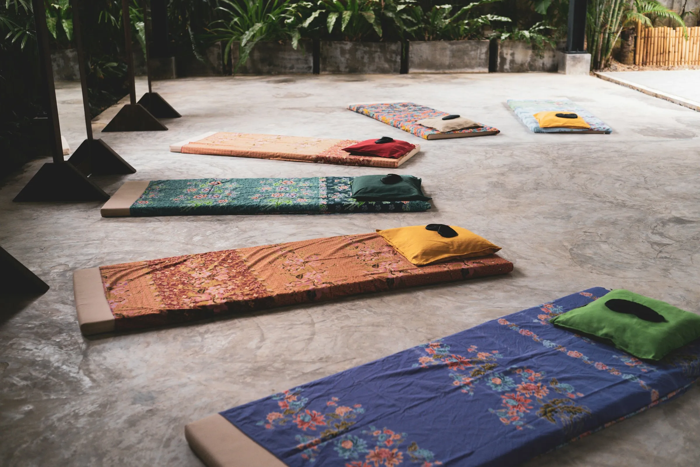 Colorful floor mats with small cushions laid out on a concrete floor in an open-air space with plants behind