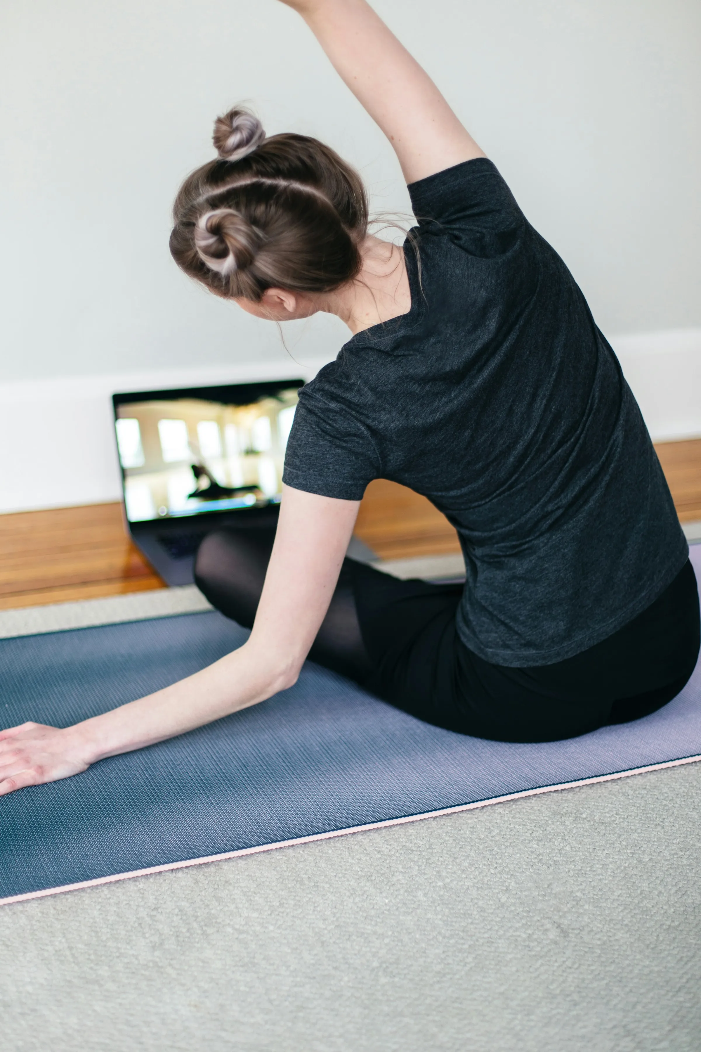 Woman in a dark t-shirt doing a seated side stretch on a blue yoga mat at home, with a laptop showing an online class beside her