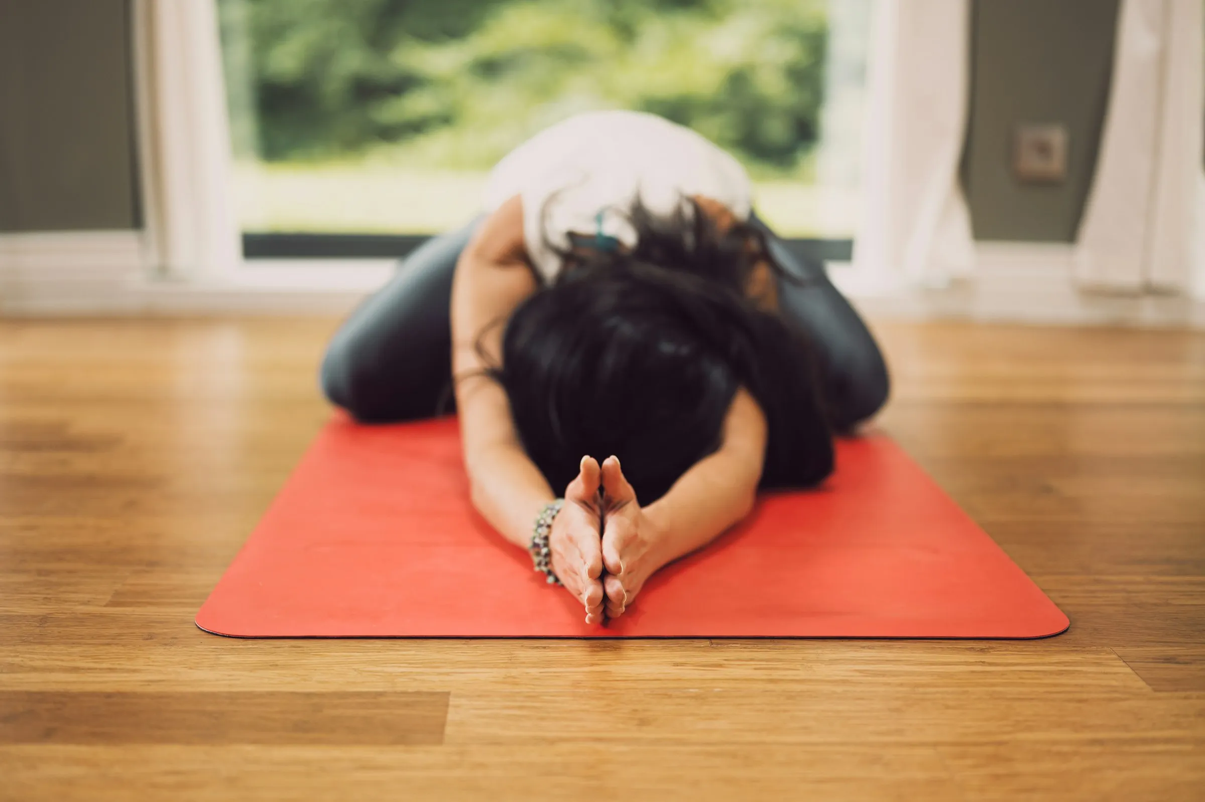 Person in child's pose on an orange yoga mat on a wooden floor in front of a bright window