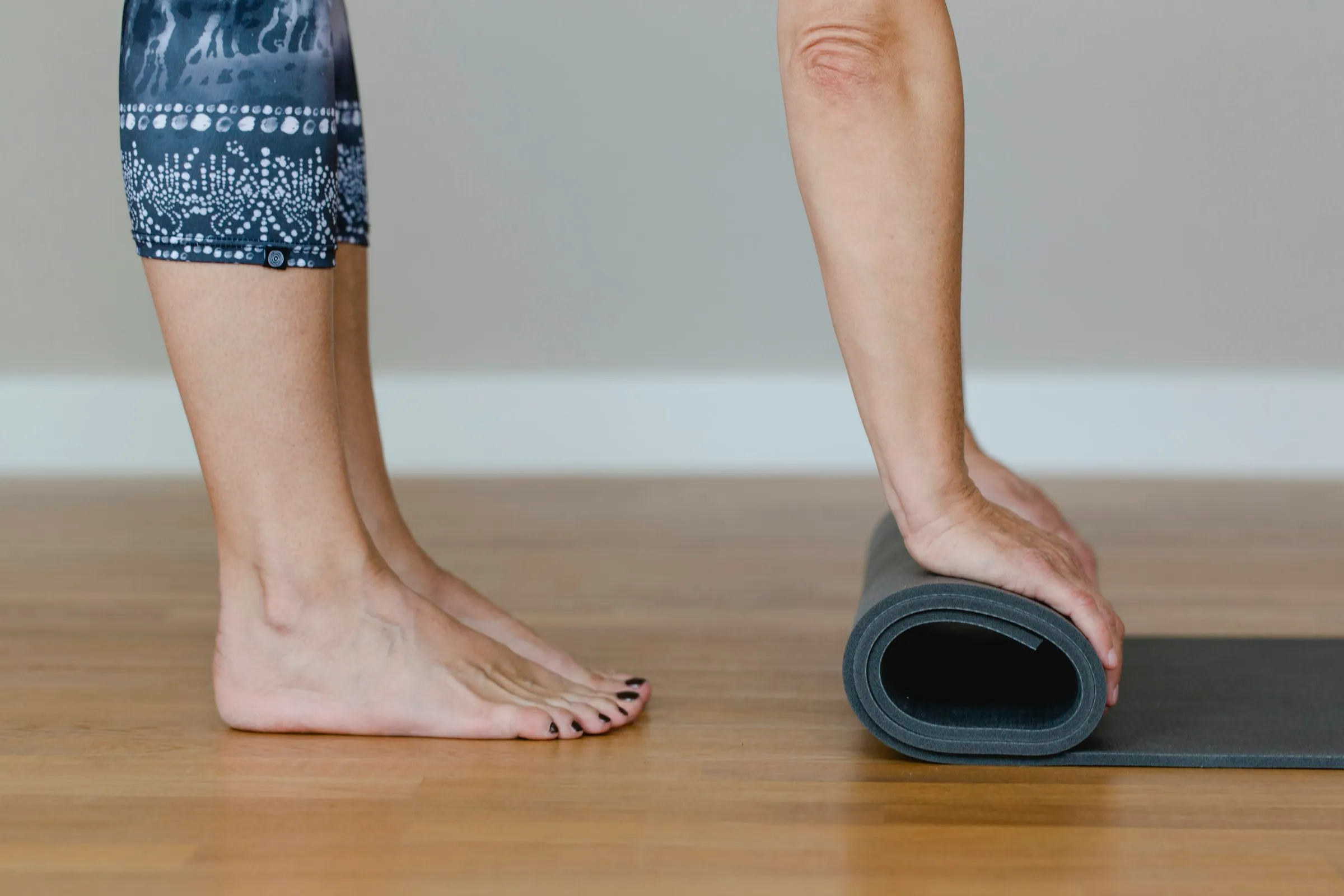 Barefoot person standing on a gray yoga mat and rolling it up on a wooden floor